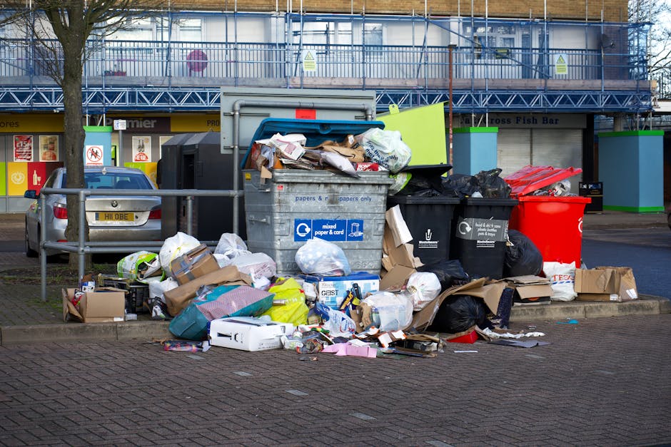 A collection of overflowing rubbish bins and scattered waste on a paved urban sidewalk, situated near a parking area with a silver car parked on the left. The waste consists of various materials inclu