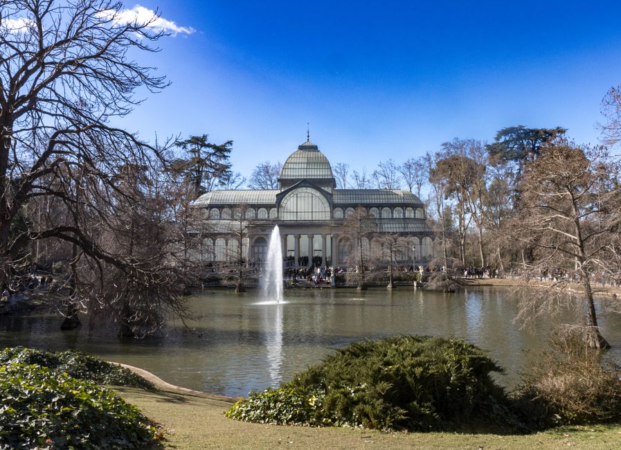 A large glass greenhouse structure situated behind a water feature in a park, with a central domed roof and extensive glass panels supported by a metal framework. In the foreground, there is a pond wi